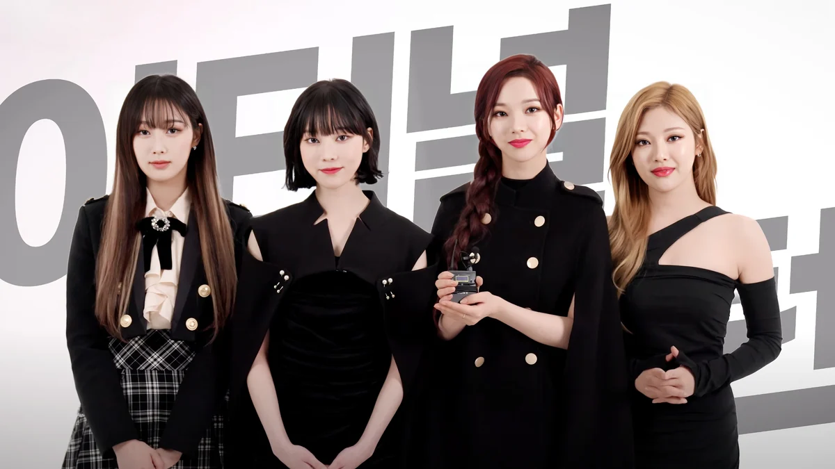 The four aespa members pose in front of a white wall; one holds a small object.