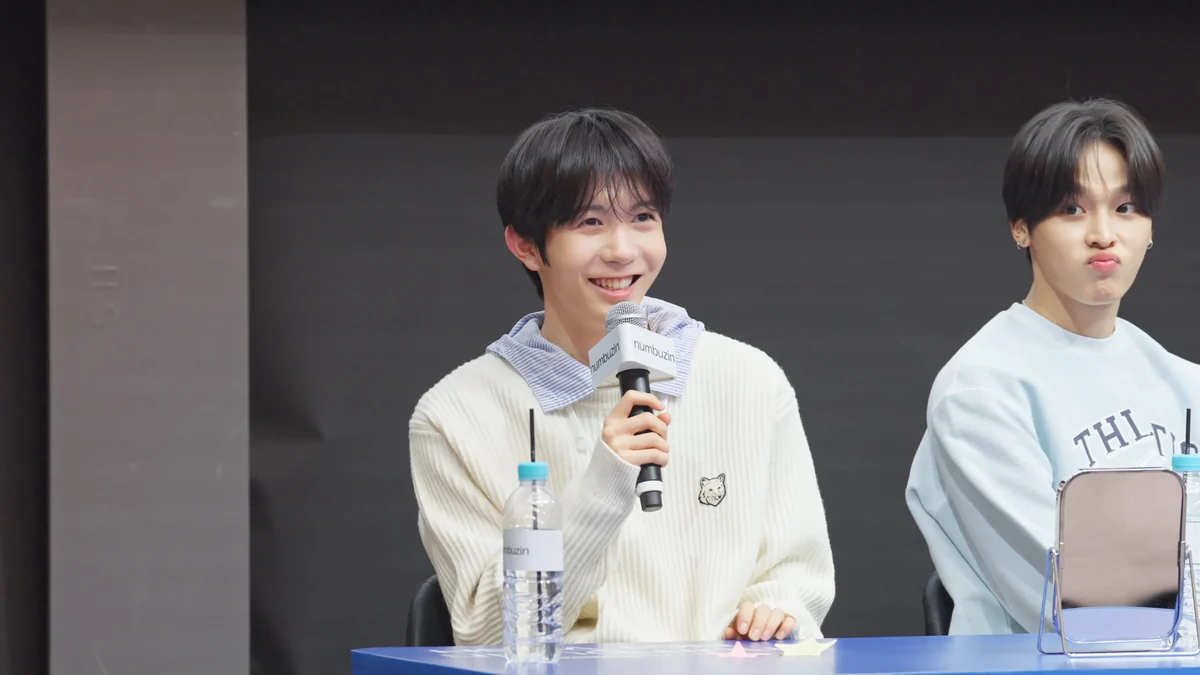 Two NCT WISH members seated at a table, one smiling while holding a microphone