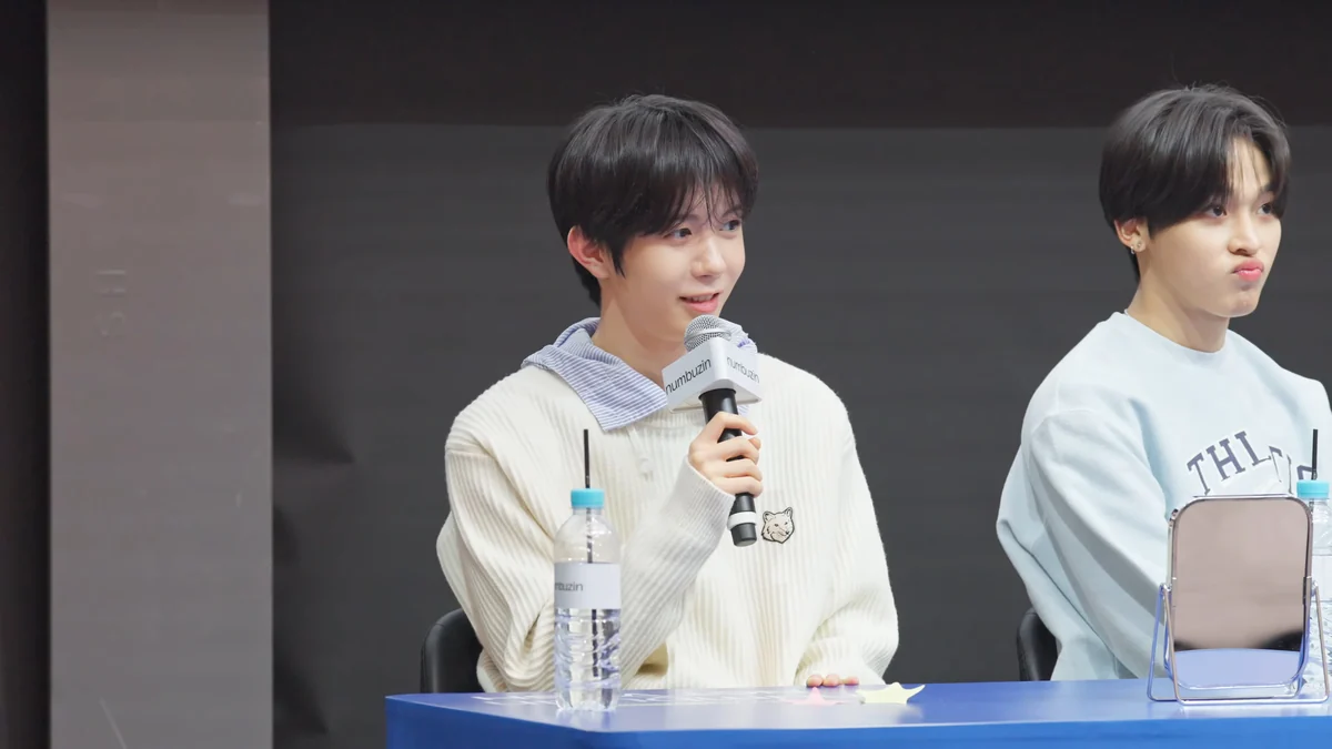 Two NCT WISH members seated at a table, one speaking into a microphone