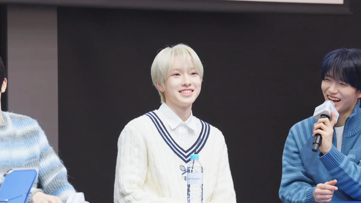 Blond NCT WISH member smiling at a table while another member holds a microphone