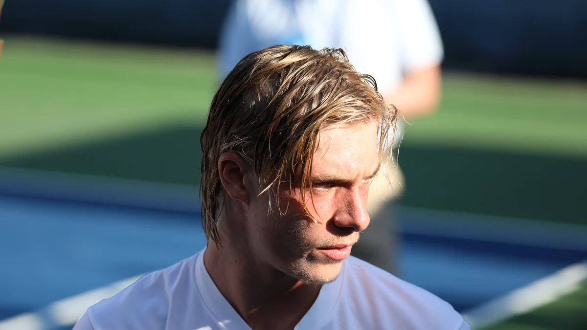 Denis Shapovalov, vêtu d’un maillot blanc, regarde sur le côté sur un court de tennis.