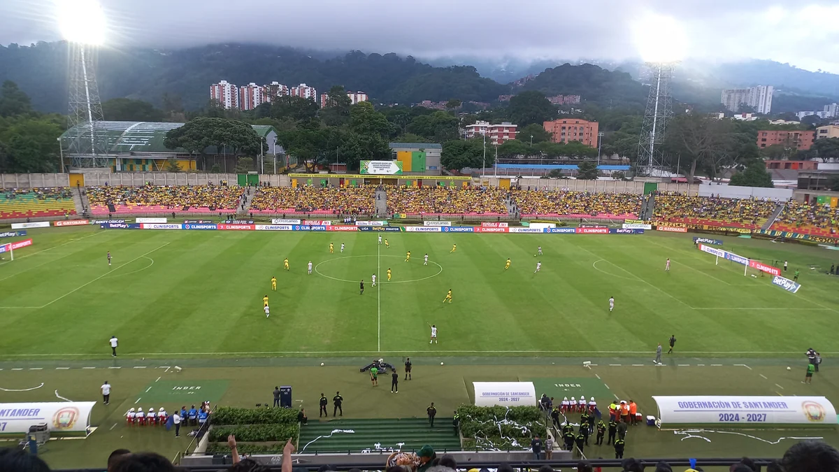 Wide view of a football match in a stadium with yellow stands and mountains behind it