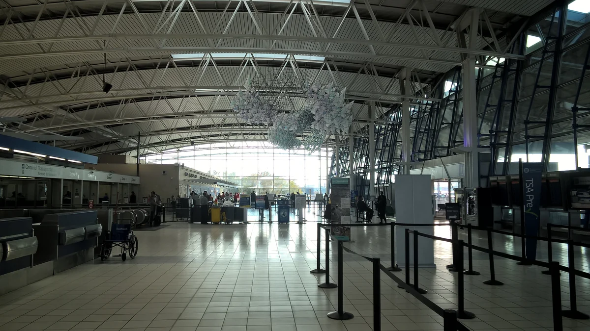 Airport hall with counters, queue barriers, and a suspended installation beneath metal roof trusses