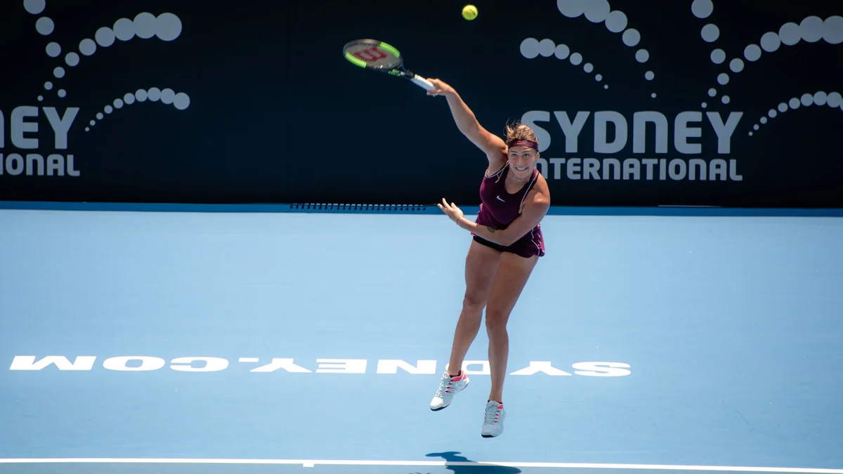 Aryna Sabalenka saca en una pista azul con la raqueta elevada sobre la cabeza.