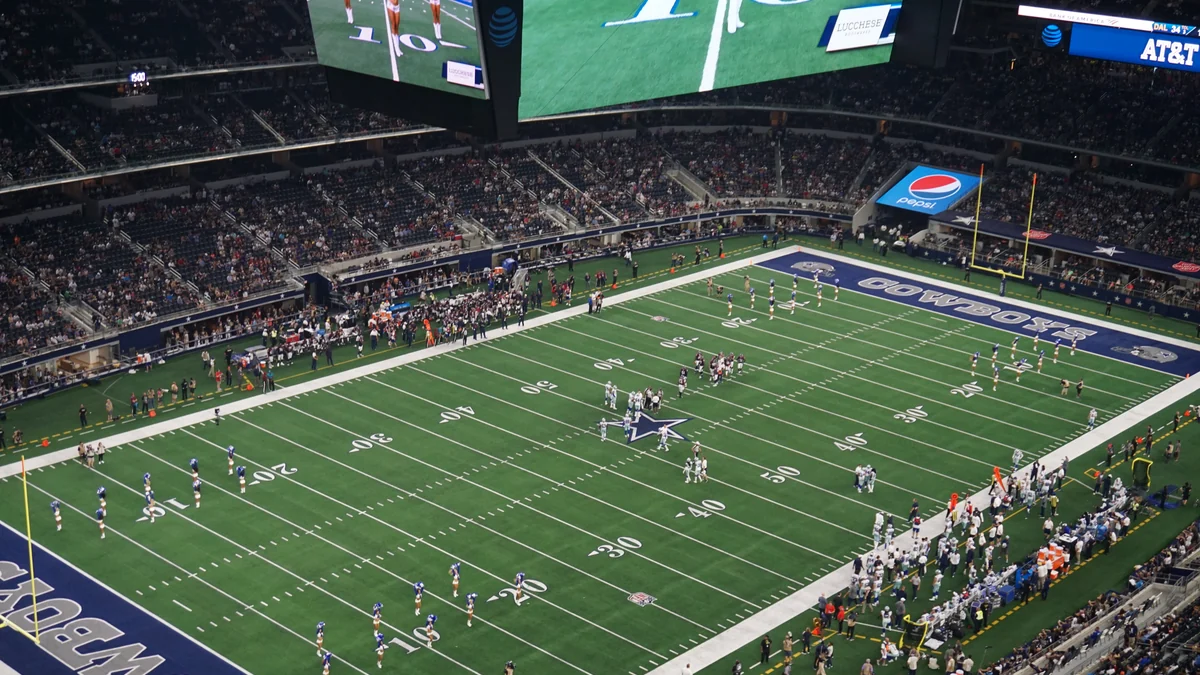 Elevated view of the football field inside AT&T Stadium, with players, occupied stands, and large suspended video screens.