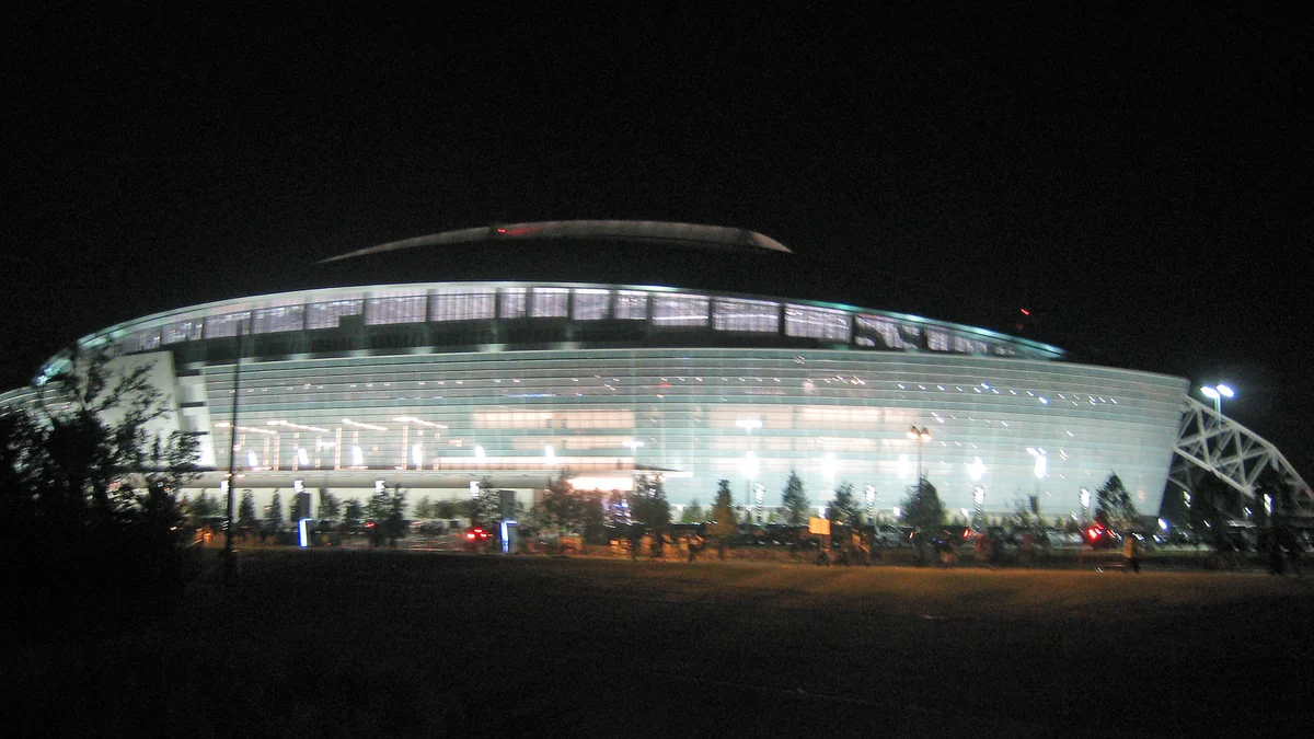 Exterior of a large stadium illuminated at night, seen behind trees and a dark foreground area.