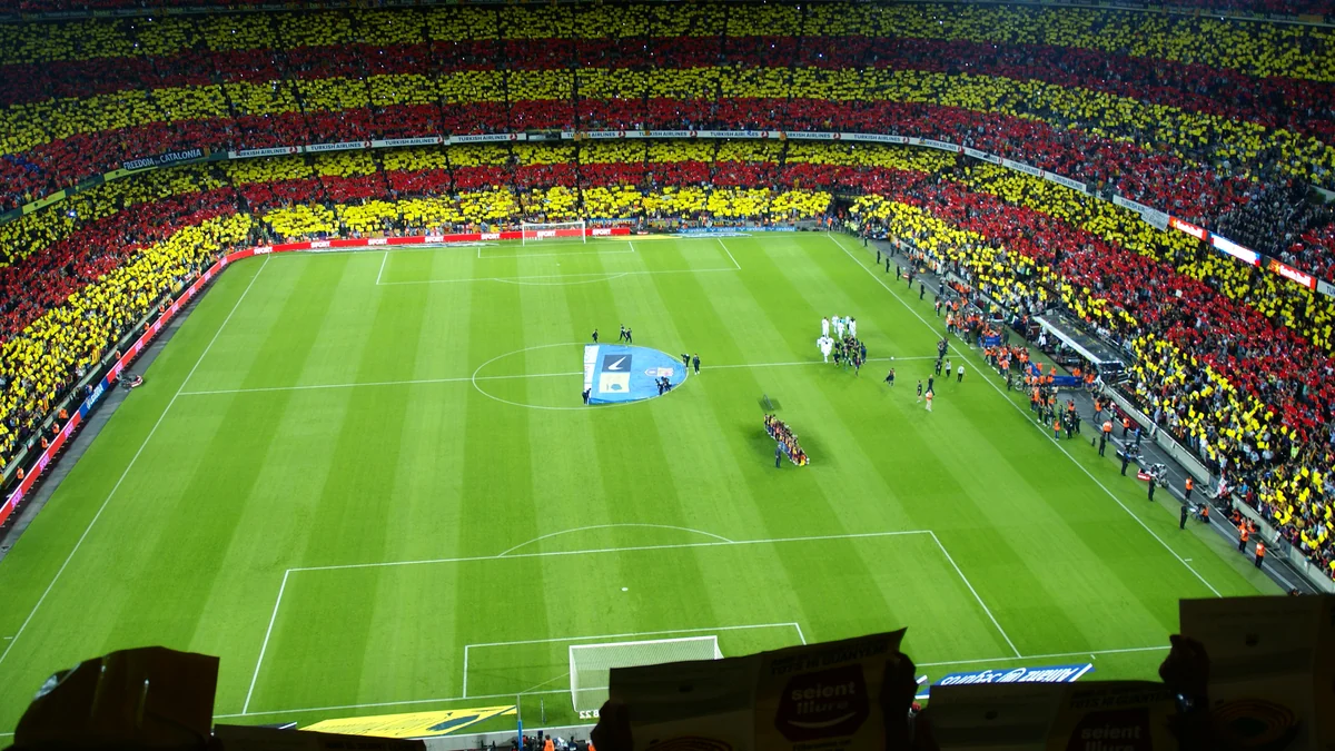 Vista ampla de um estádio de futebol lotado, com mosaico vermelho e amarelo nas arquibancadas e jogadores reunidos no campo.