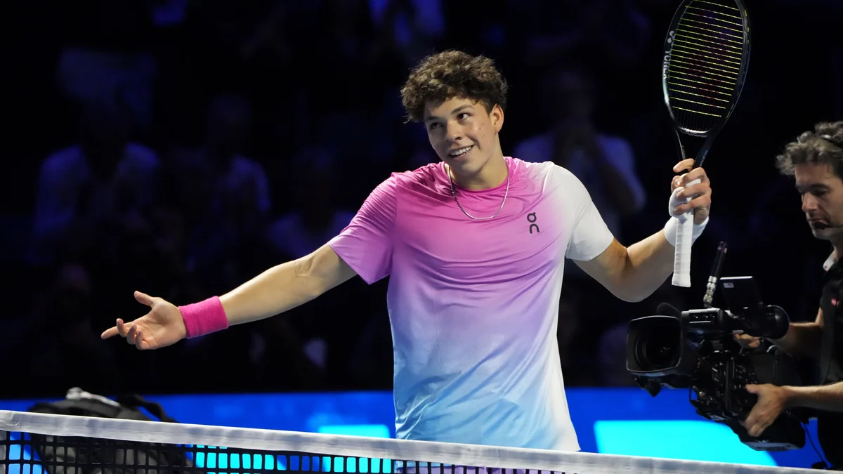 Ben Shelton sonríe con la raqueta levantada frente a la red en una pista de tenis cubierta.