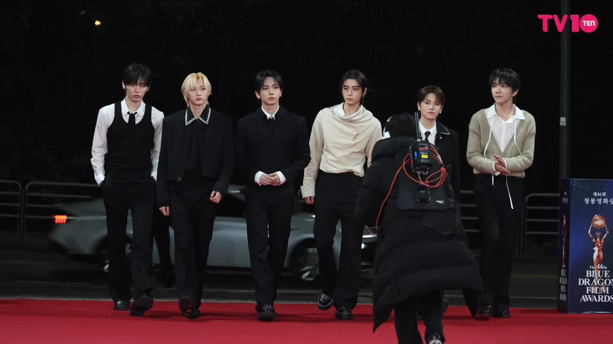 Six BOYNEXTDOOR members on a red carpet with a camera operator in the foreground