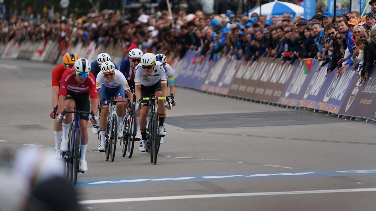 A group of cyclists crosses the finishing straight beside barriers lined with spectators