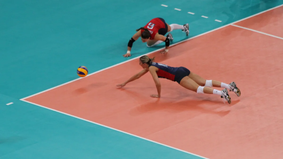 Two volleyball players dive toward the ball near the sideline.