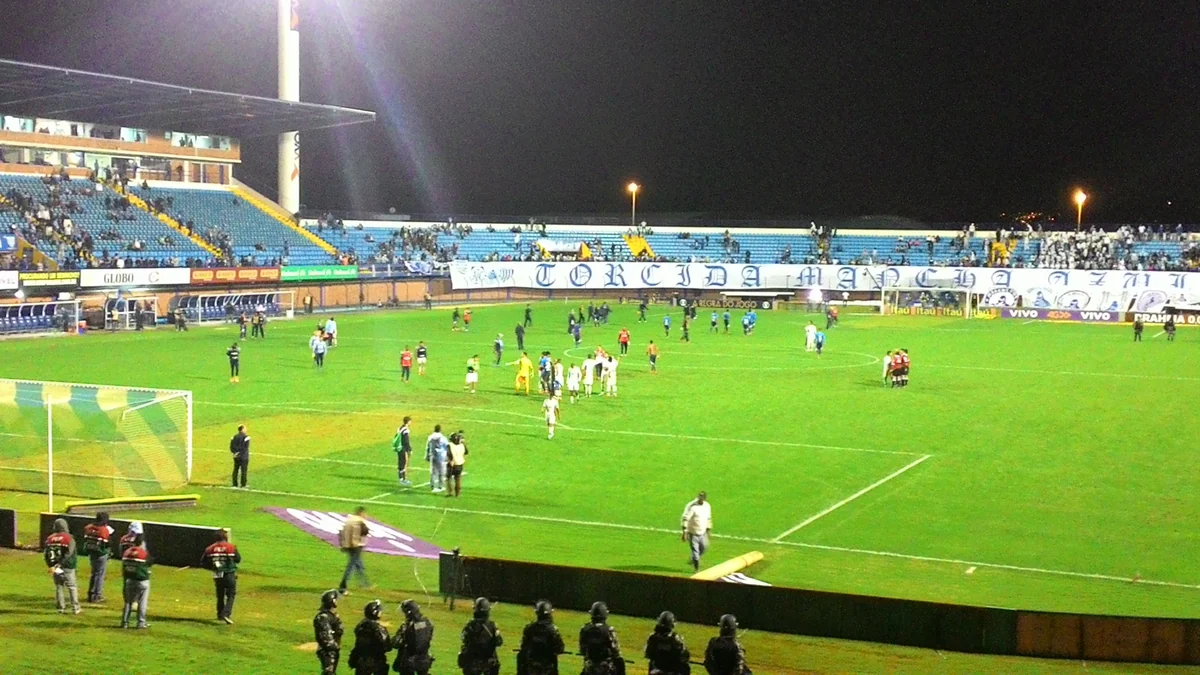 Vista ampla de campo de futebol iluminado à noite, com jogadores e policiais junto à arquibancada