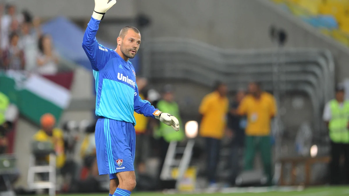 Goleiro de uniforme azul levanta uma das mãos no gramado