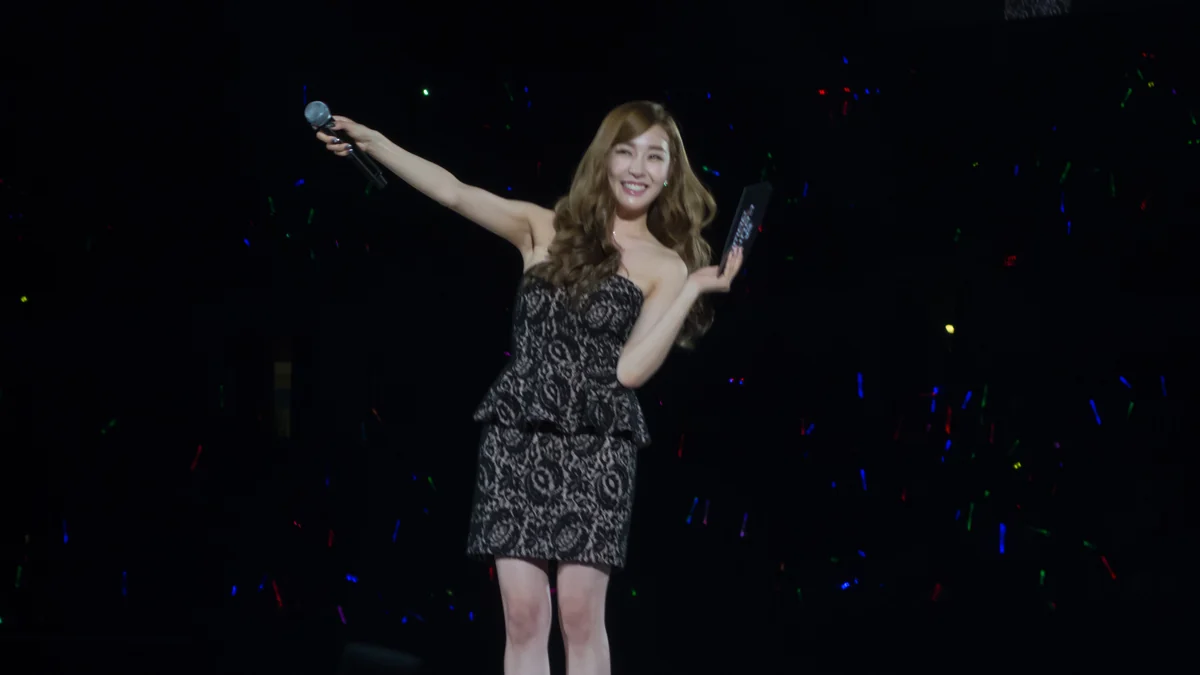 Tiffany Young sorri no palco com um microfone e cartões nas mãos diante de luzes coloridas do público.