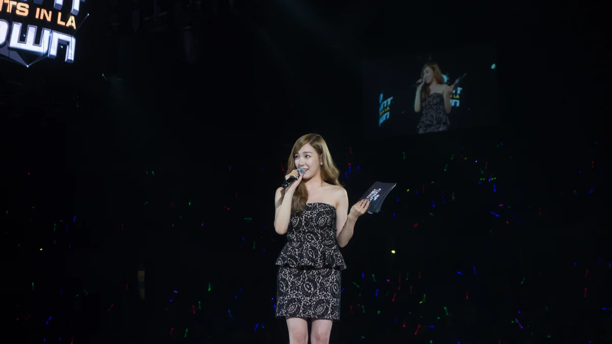 Tiffany Young fala ao microfone no palco enquanto segura cartões, com uma tela e luzes do público ao fundo.