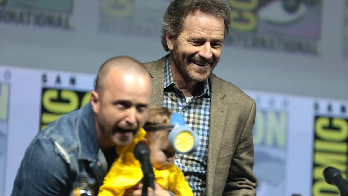 Bryan Cranston smiling behind Aaron Paul, who holds a child in front of a microphone