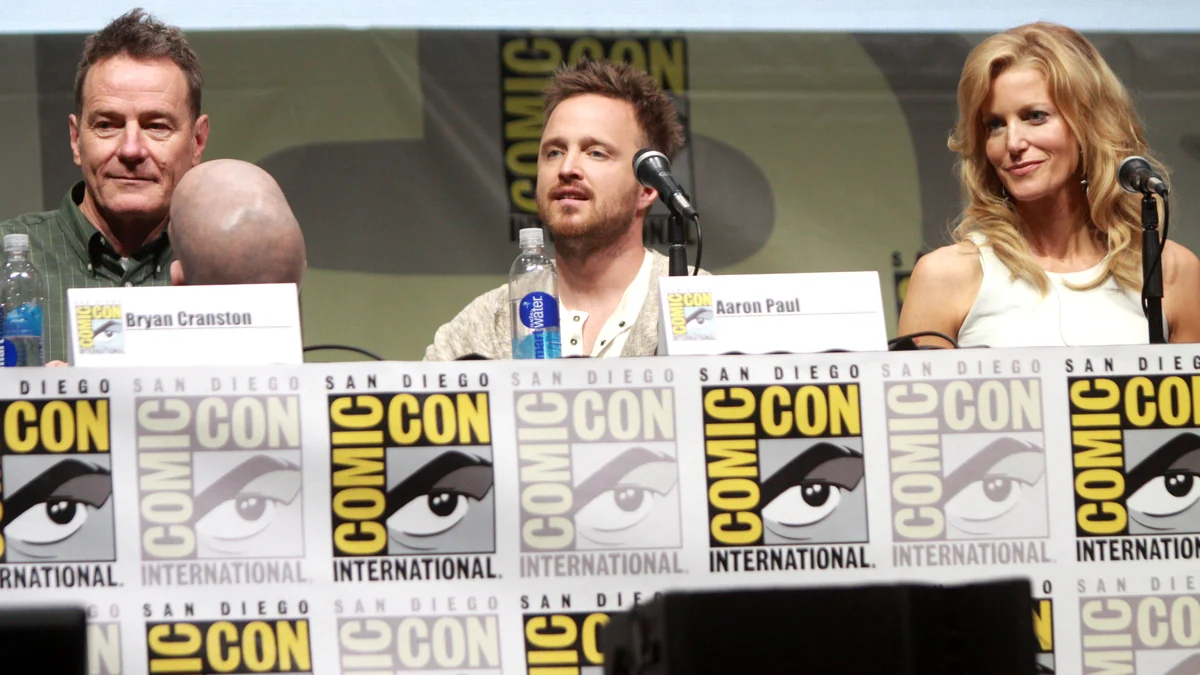 Bryan Cranston, Aaron Paul, and Anna Gunn seated behind a table decorated with Comic-Con signs