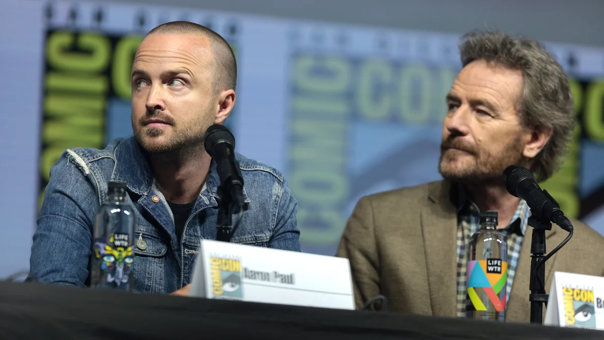 Aaron Paul and Bryan Cranston looking to the side during a panel, with microphones in front of them