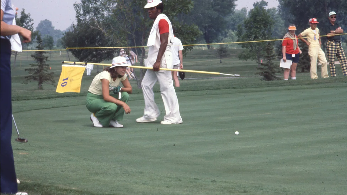 Une golfeuse accroupie observe une balle sur le green tandis qu’un cadet passe avec le drapeau