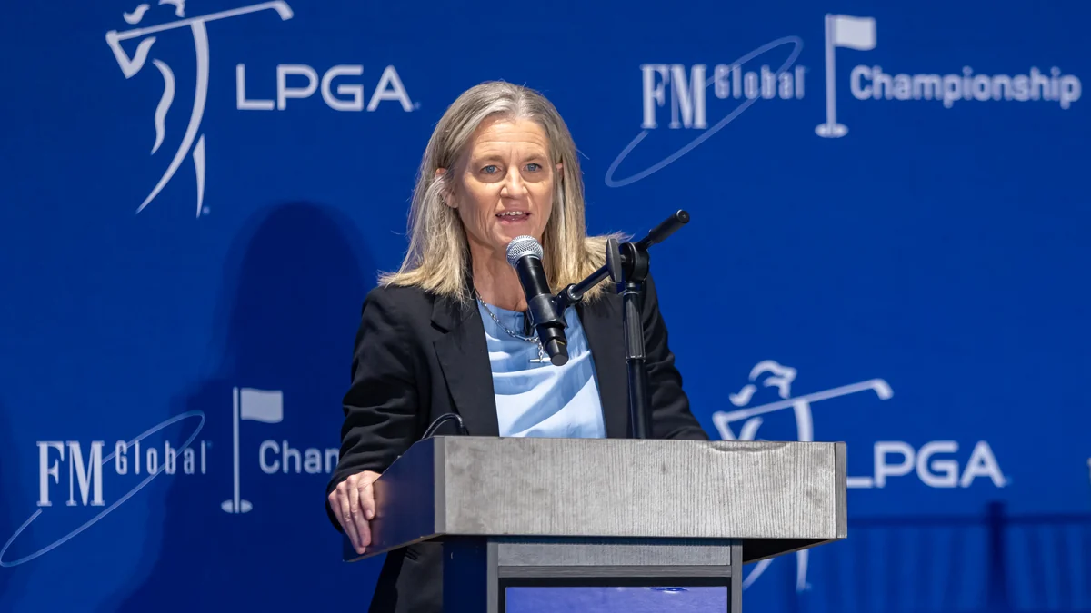Mujer habla ante un atril frente a un panel azul con logotipos de la LPGA y el FM Global Championship