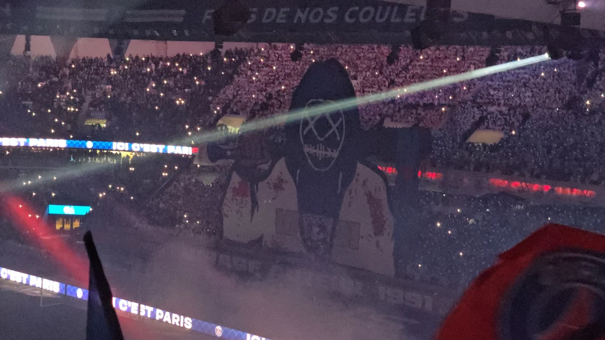 A large tifo depicting a hooded figure covers a stand amid lights and smoke.