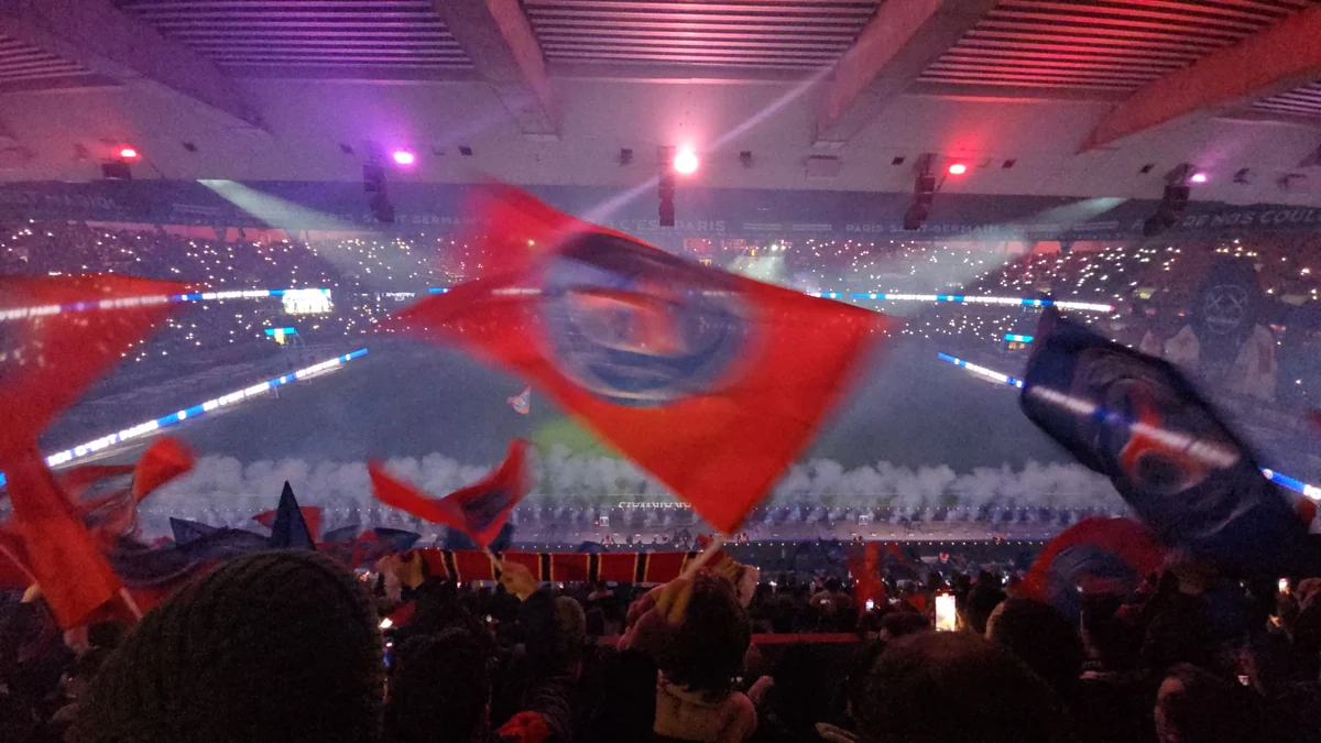 Supporters wave large red and blue PSG flags in a lit stadium stand.