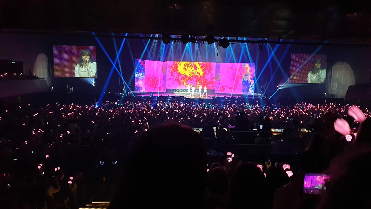 Colorful stage at the far end of a dark arena, with the audience and a phone filming in the foreground