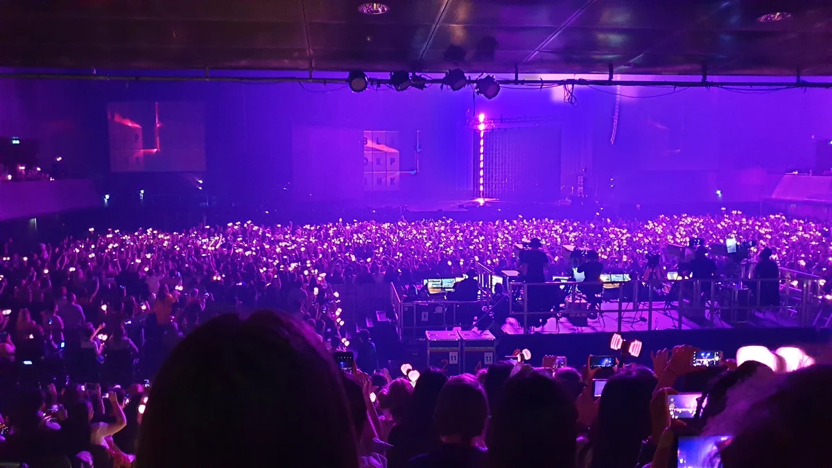 Packed arena under purple light, with phones and light sticks raised toward the stage