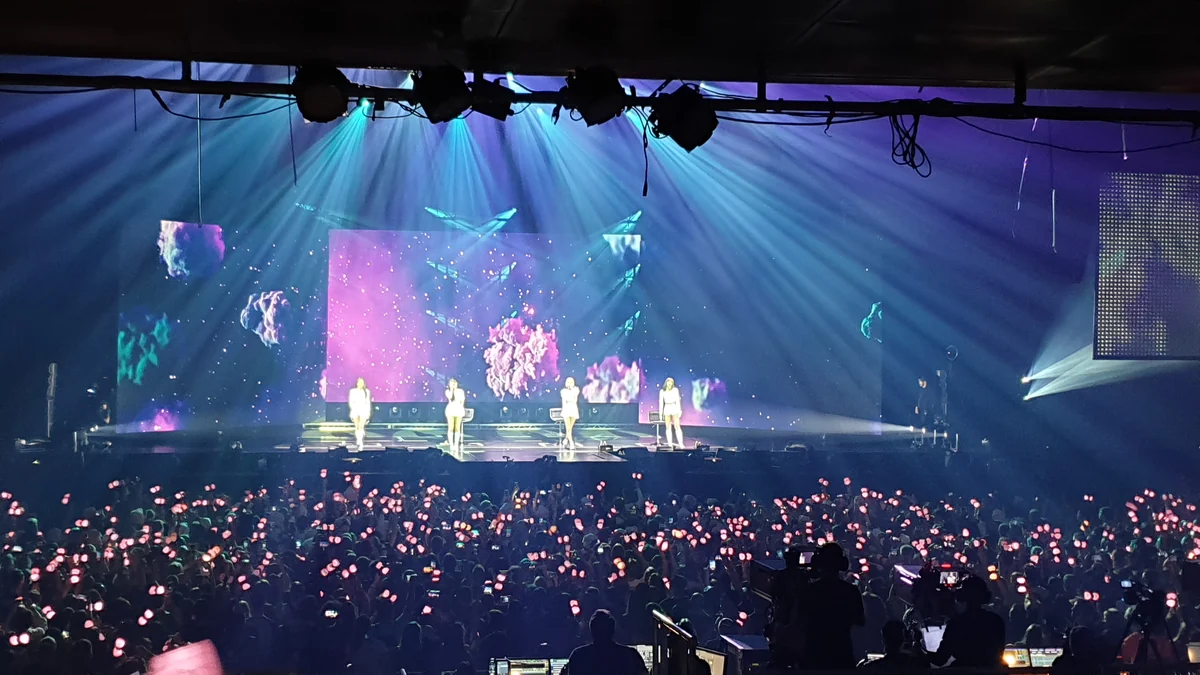 Four performers on a lit stage before an audience holding light sticks and phones