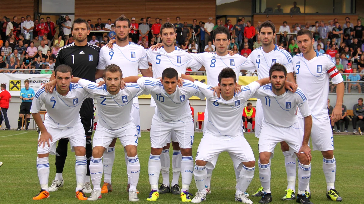 Onze jogadores da seleção grega sub-21 posam no gramado, usando uniformes brancos e um uniforme preto de goleiro.