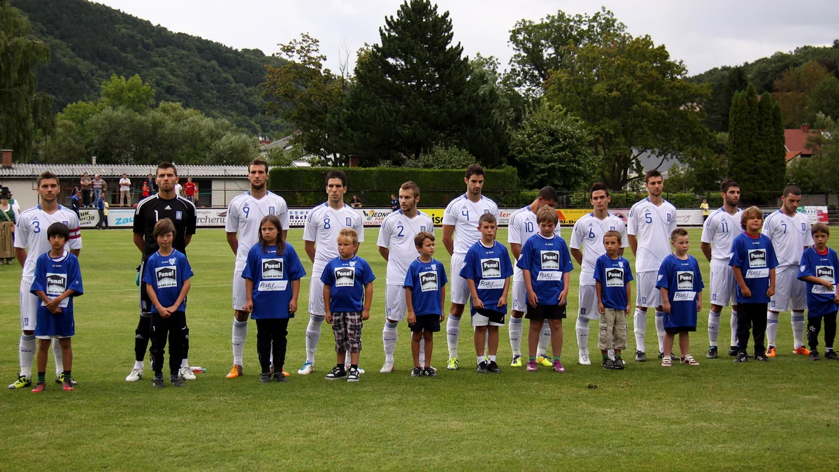 Jogadores da seleção grega sub-21 formam uma fila atrás de crianças vestidas com camisetas azuis em um campo cercado por árvores.