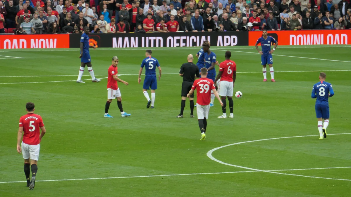 Jugadores del Manchester United, de rojo, y del Chelsea, de azul, colocados alrededor del árbitro sobre el césped.