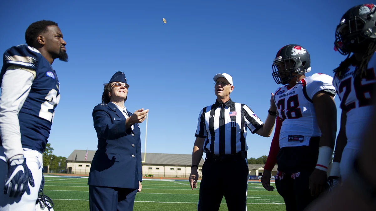 Military officer flips a coin beside a referee and football players on the field
