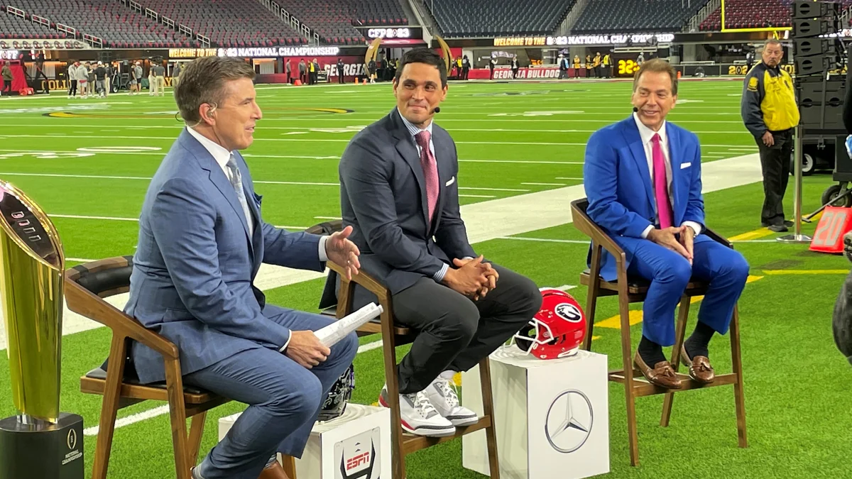 Três comentaristas sentados à beira do campo ao lado do troféu do College Football Playoff