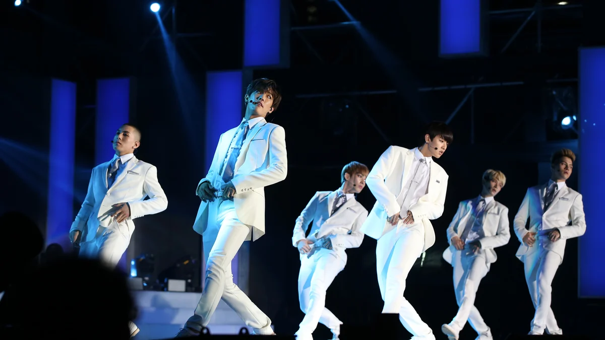 Six group members dance onstage in white suits under blue lighting.