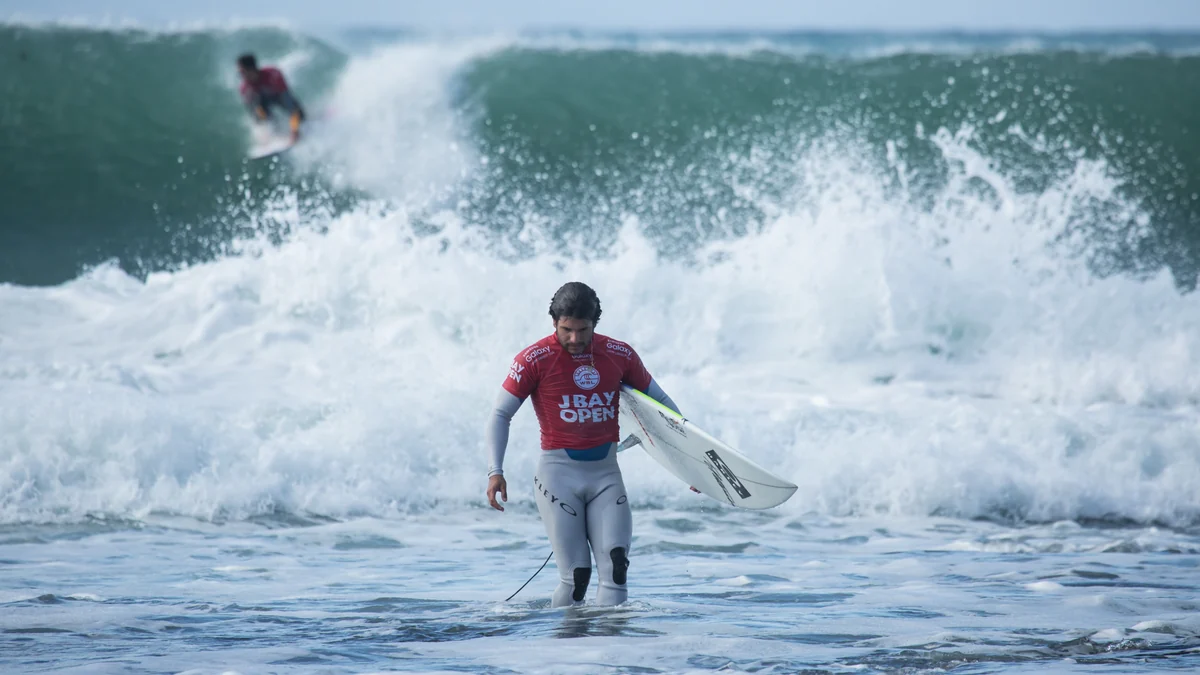 Surfista con licra roja camina por el agua con su tabla mientras otro atleta surfea al fondo