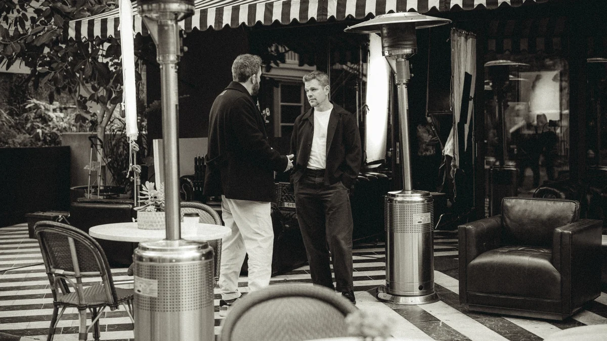 Matt Damon conversa con Ben Affleck en una terraza con mesas, sillas y calentadores