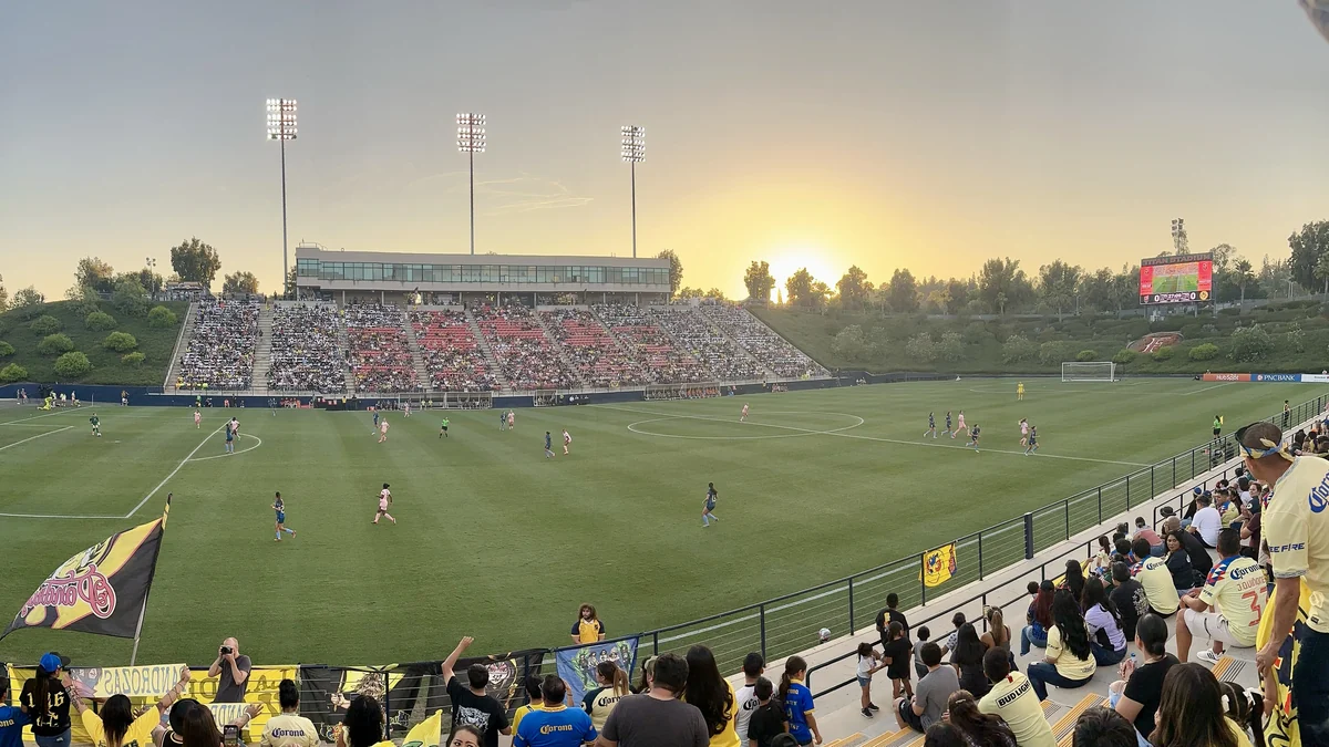 Football pitch during a women’s match, with players in pink and dark blue and stands in the background