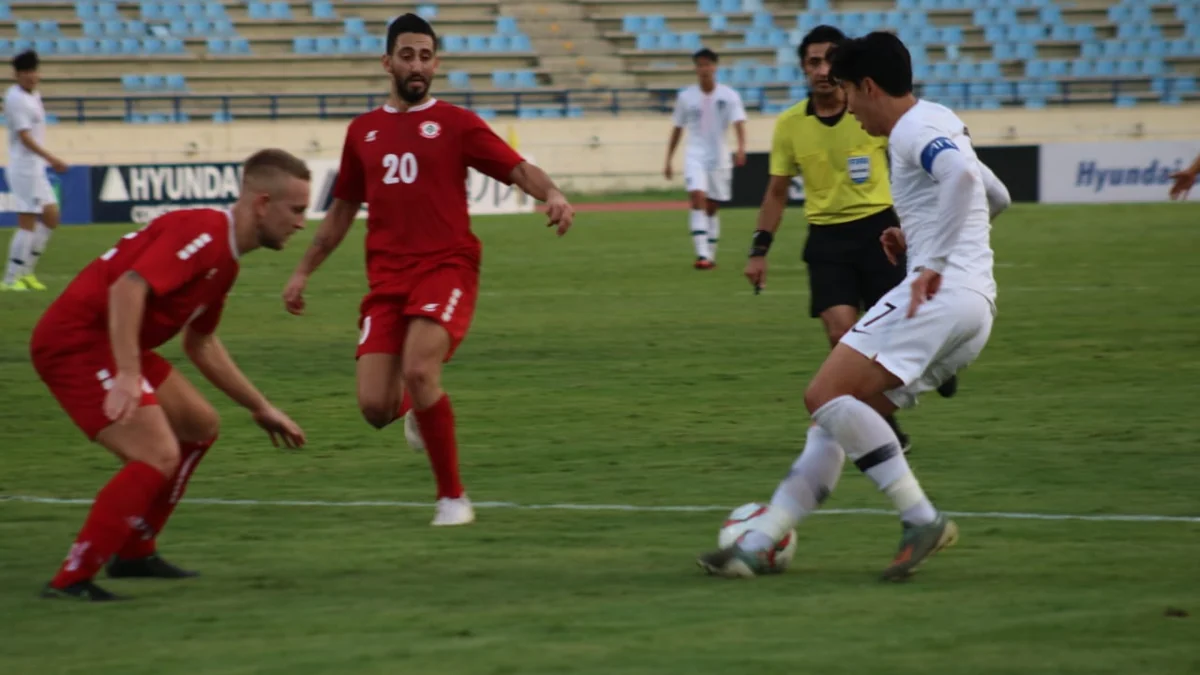 A South Korea player in white controls the ball as opponents in red close in and the referee watches.