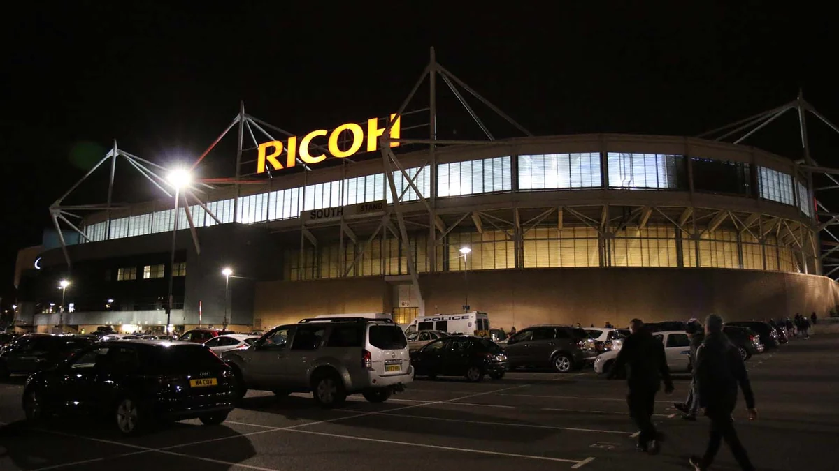 Coventry stadium exterior at night with the illuminated Ricoh sign and cars in the parking area
