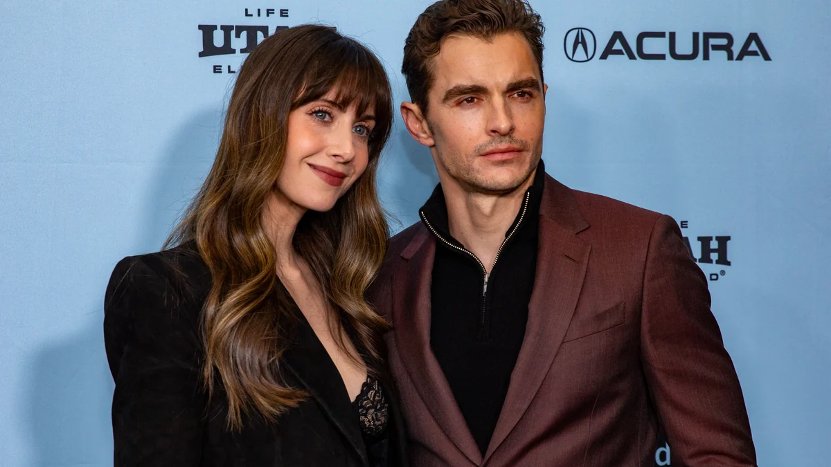 Alison Brie e Dave Franco posam juntos diante de um painel azul de evento.