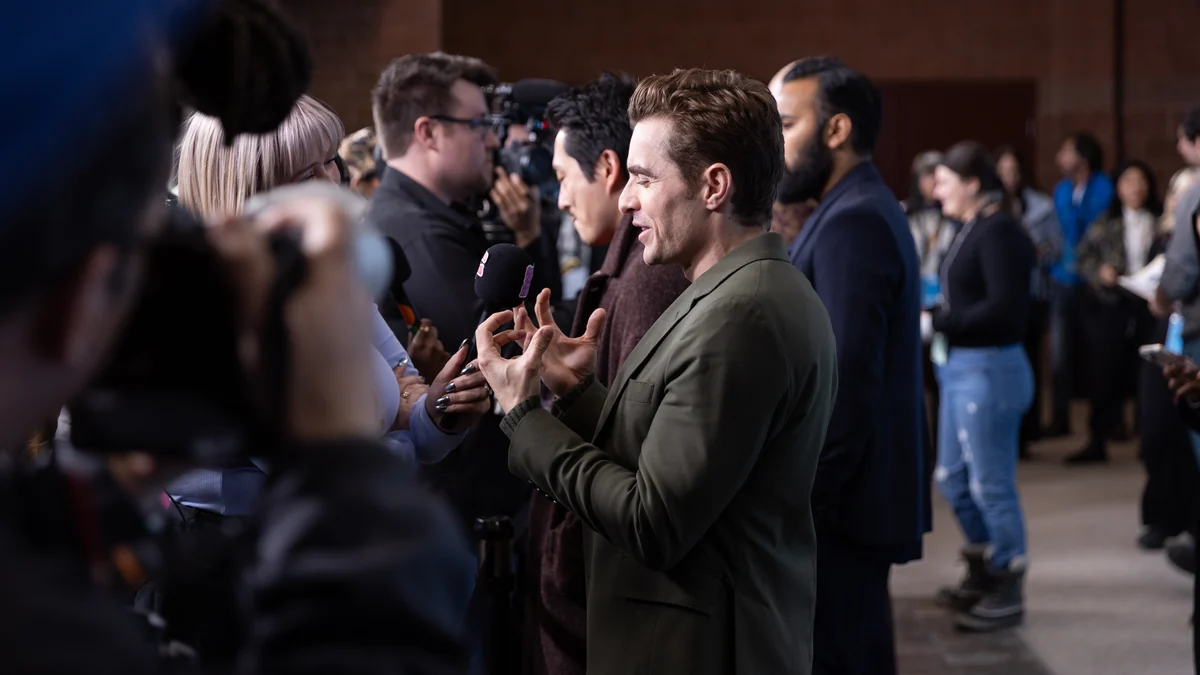 Dave Franco, de perfil, fala com jornalistas enquanto gesticula em uma área de imprensa.