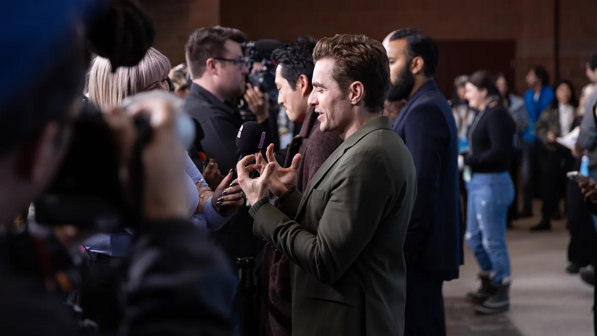 Dave Franco, de perfil, fala com jornalistas enquanto gesticula em uma área de imprensa.