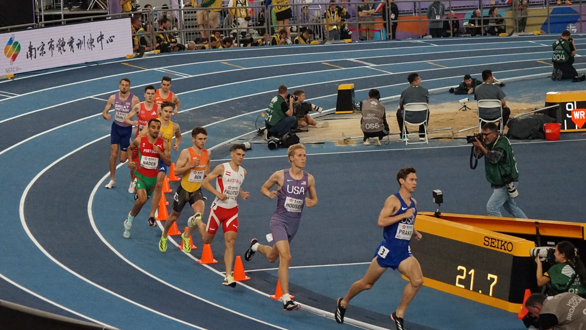 A group of runners rounds the bend on a blue track during a men’s middle-distance race.