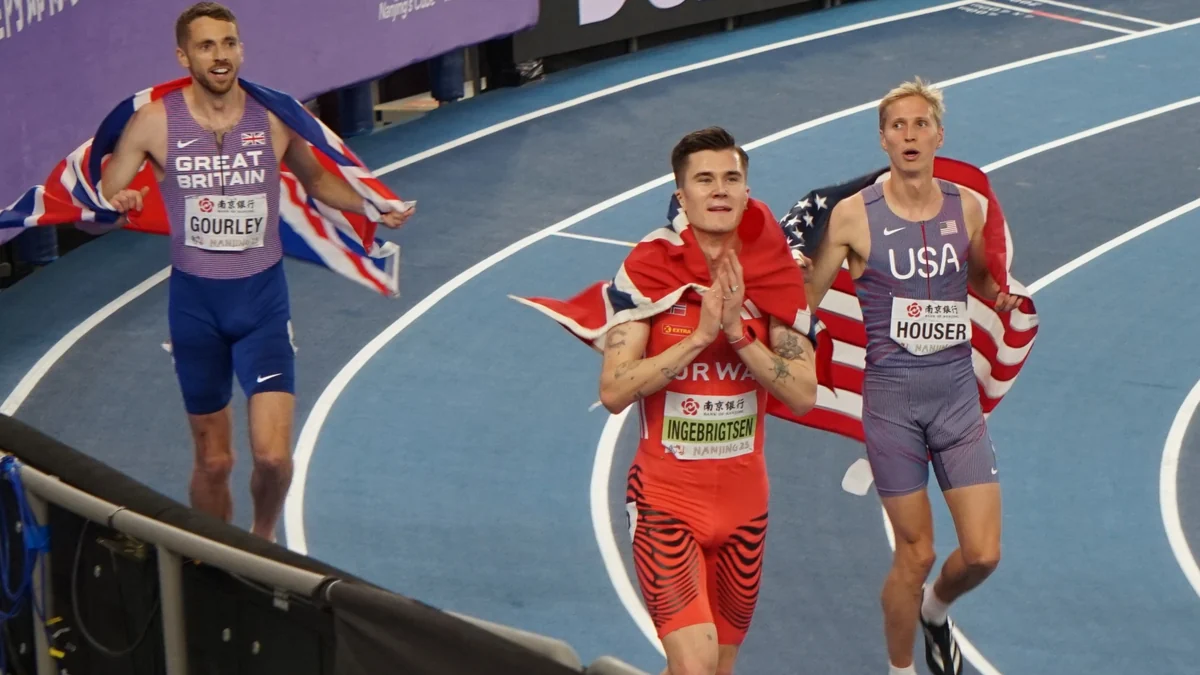 Jakob Ingebrigtsen, with a Norwegian flag over his shoulders, walks beside two runners on the track.