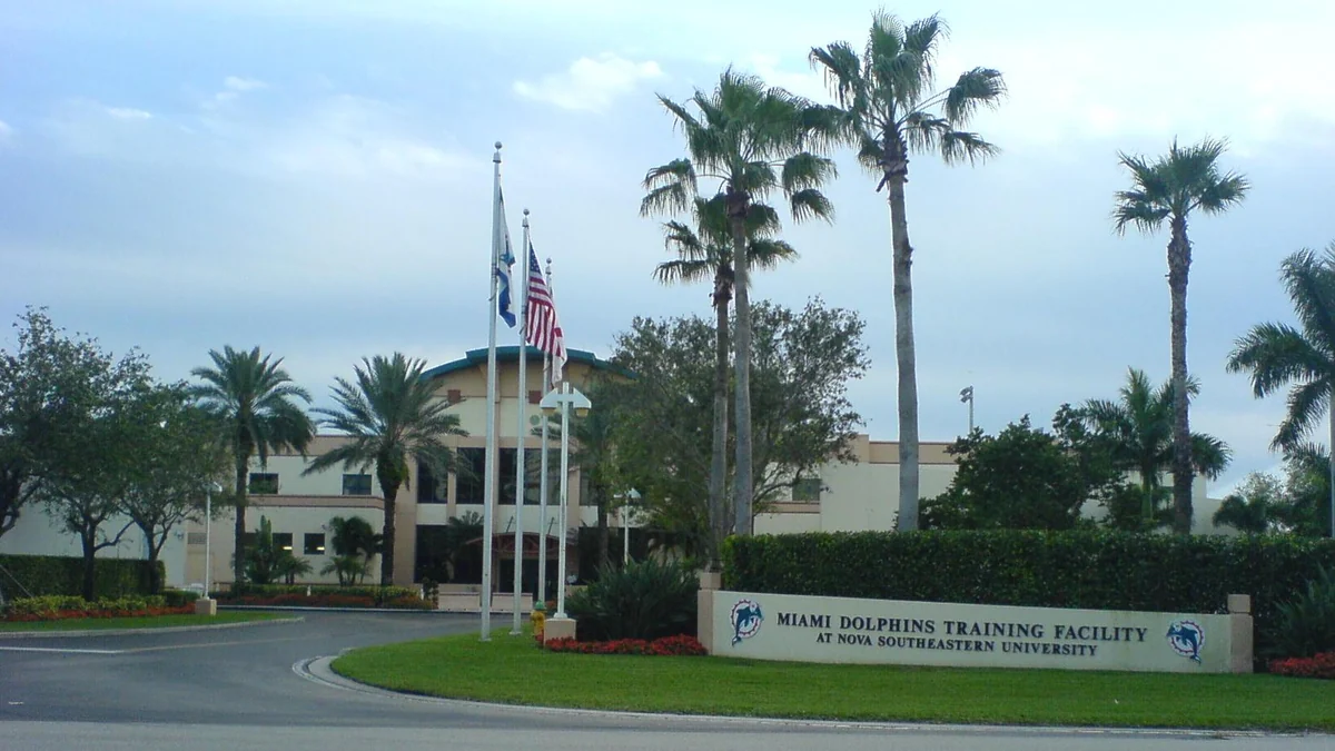 Entrada rodeada de palmeras, con banderas y un letrero del centro de entrenamiento de los Miami Dolphins en Nova Southeastern University.