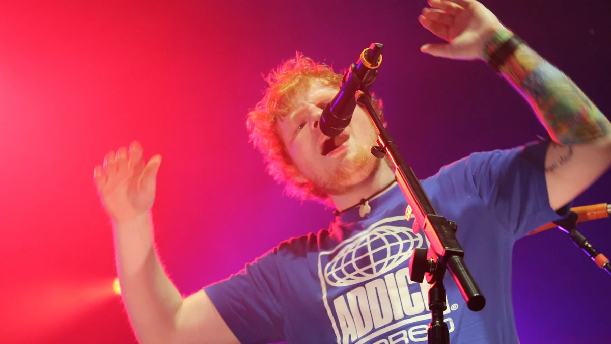 Ed Sheeran canta ao microfone com os braços erguidos sob luzes vermelhas e roxas