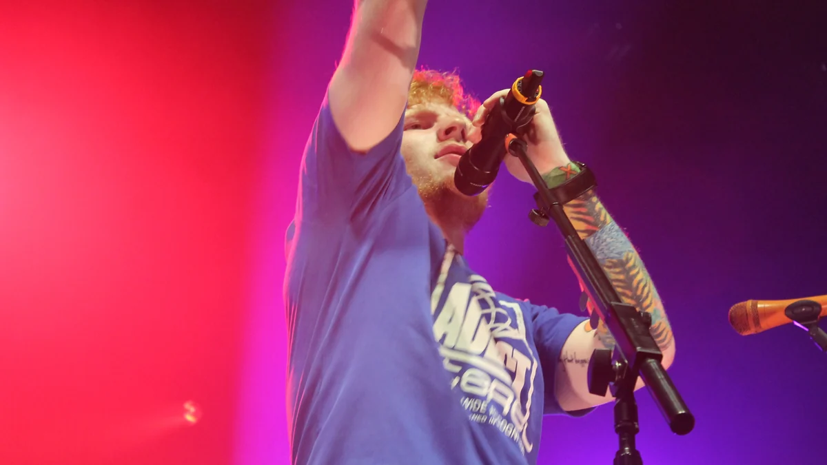 Ed Sheeran canta ao microfone com um braço levantado, iluminado em vermelho e roxo