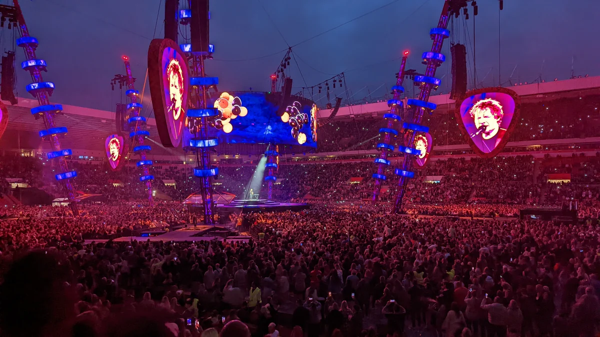 Palco de Ed Sheeran em um estádio lotado, com grandes telas e iluminação azul e vermelha