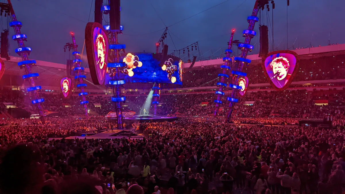 Palco de Ed Sheeran em um estádio lotado, com grandes telas e iluminação azul e vermelha